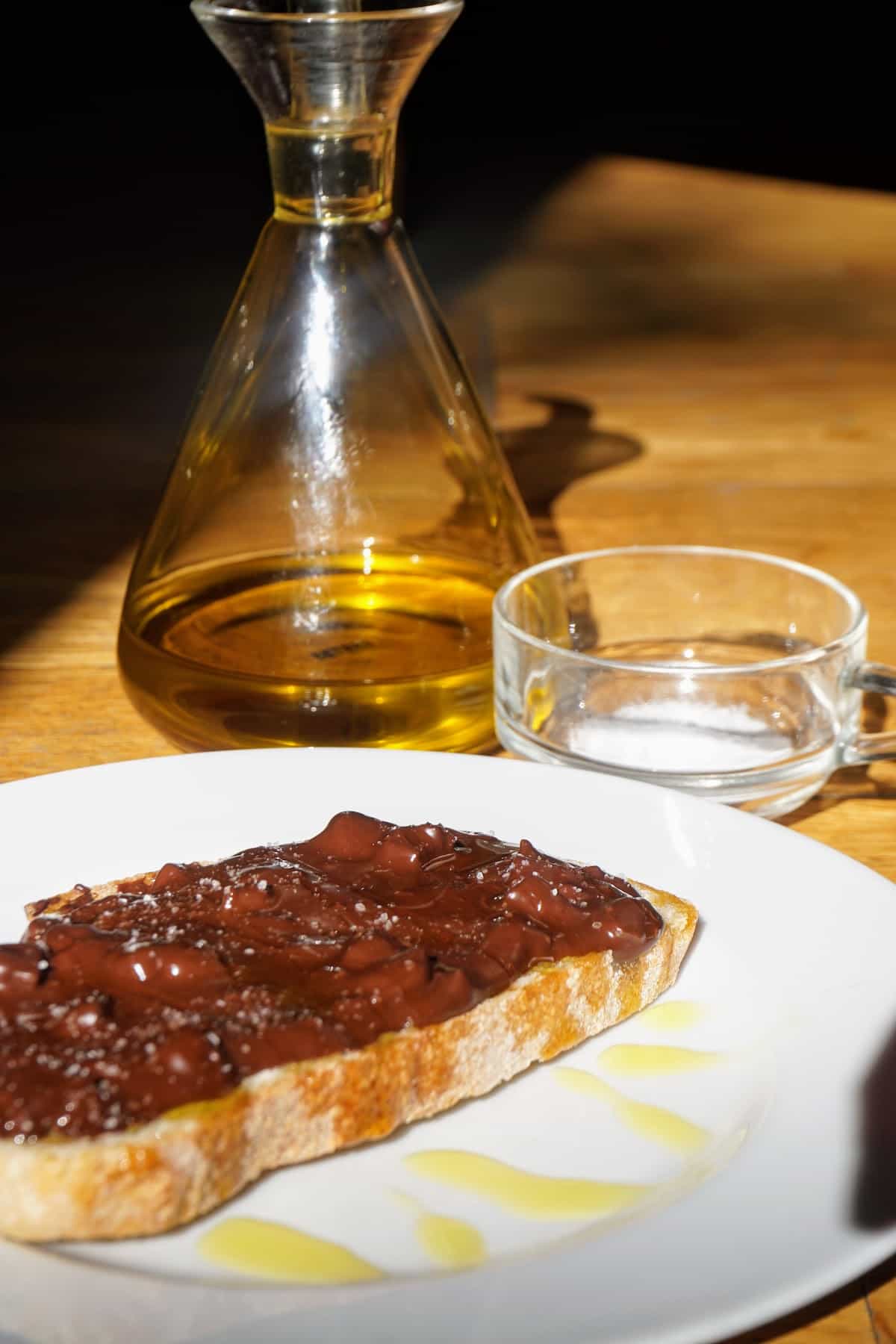 Slice of toasted bread topped with chocolate and olive oil, with containers of olive oil and salt behind the plate.