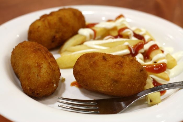 A plate with three oblong croquettes, French fries with ketchup and mayonnaise, and a fork.