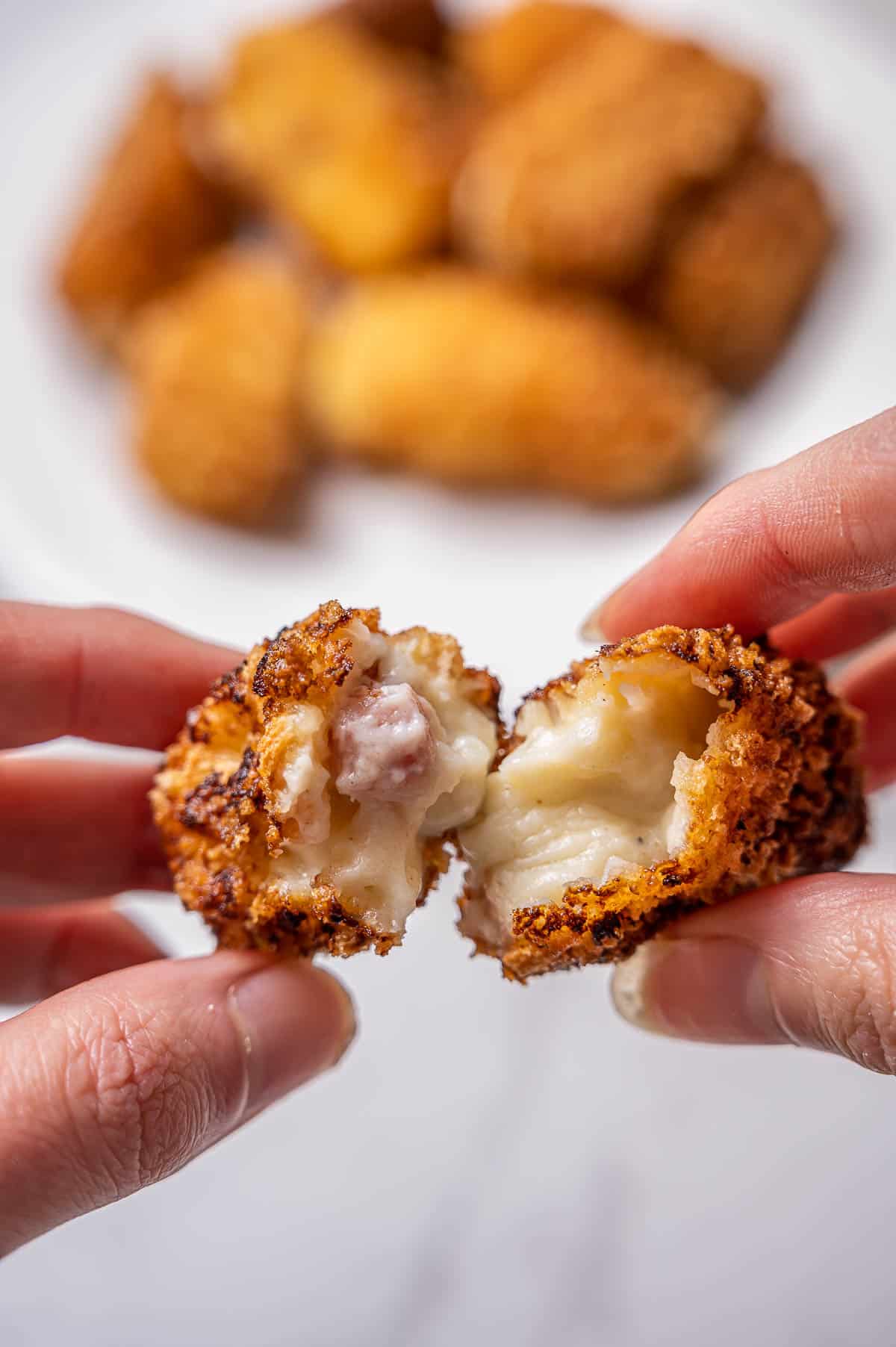 Close up of hands holding an open freshly fried Spanish ham croquette. Plate of croquetas in the background.