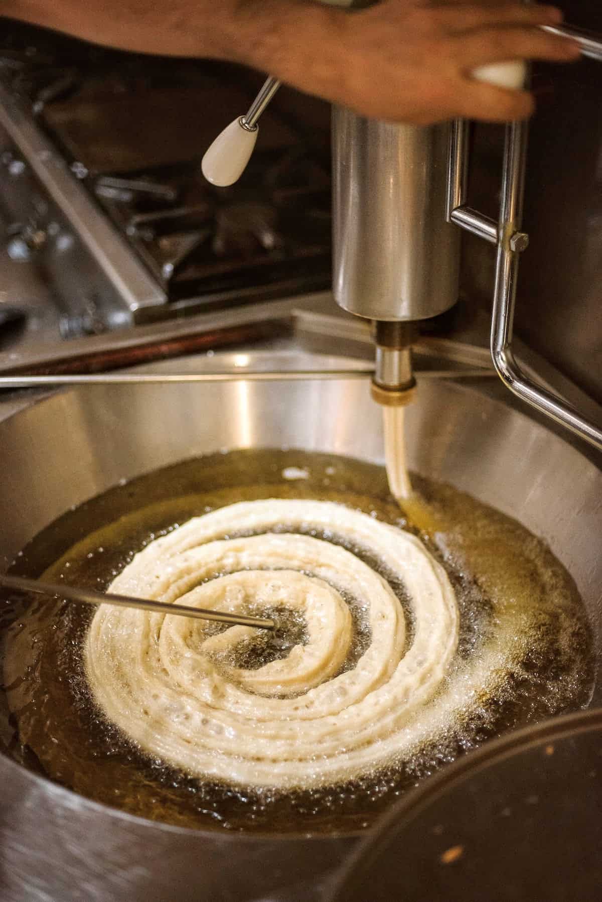 Spiral shaped churro dough being fried in a metal vat of olive oil.