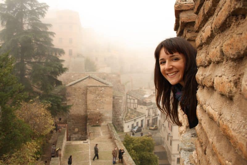 Smiling woman leans out of a stone structure with fog and old buildings in the background.