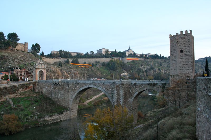 A wide view of the Alcántara Bridge, with stone arches and a tower at one end.