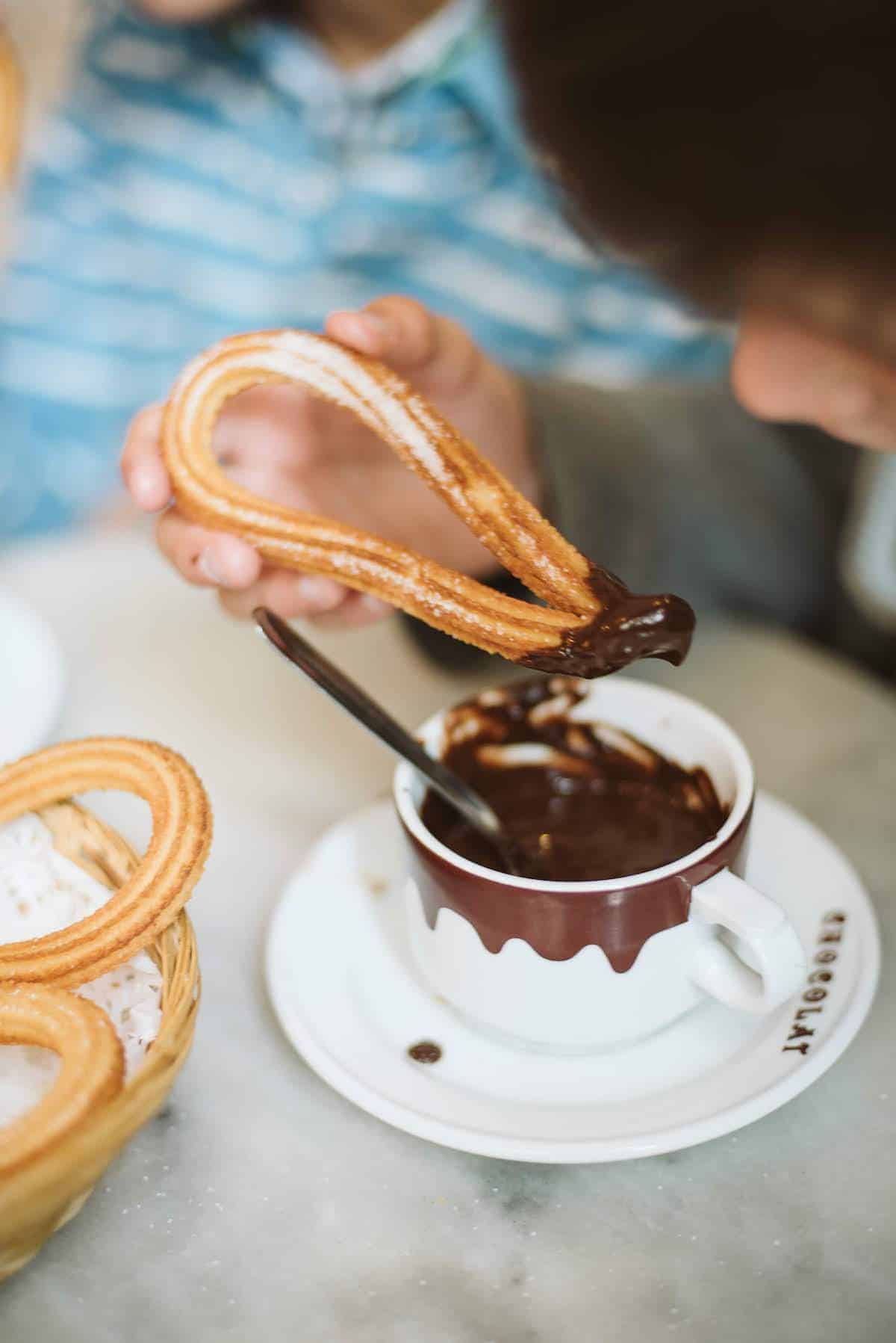 Close-up of a thin, looped churro being dipped into a mug of thick Spanish hot chocolate.