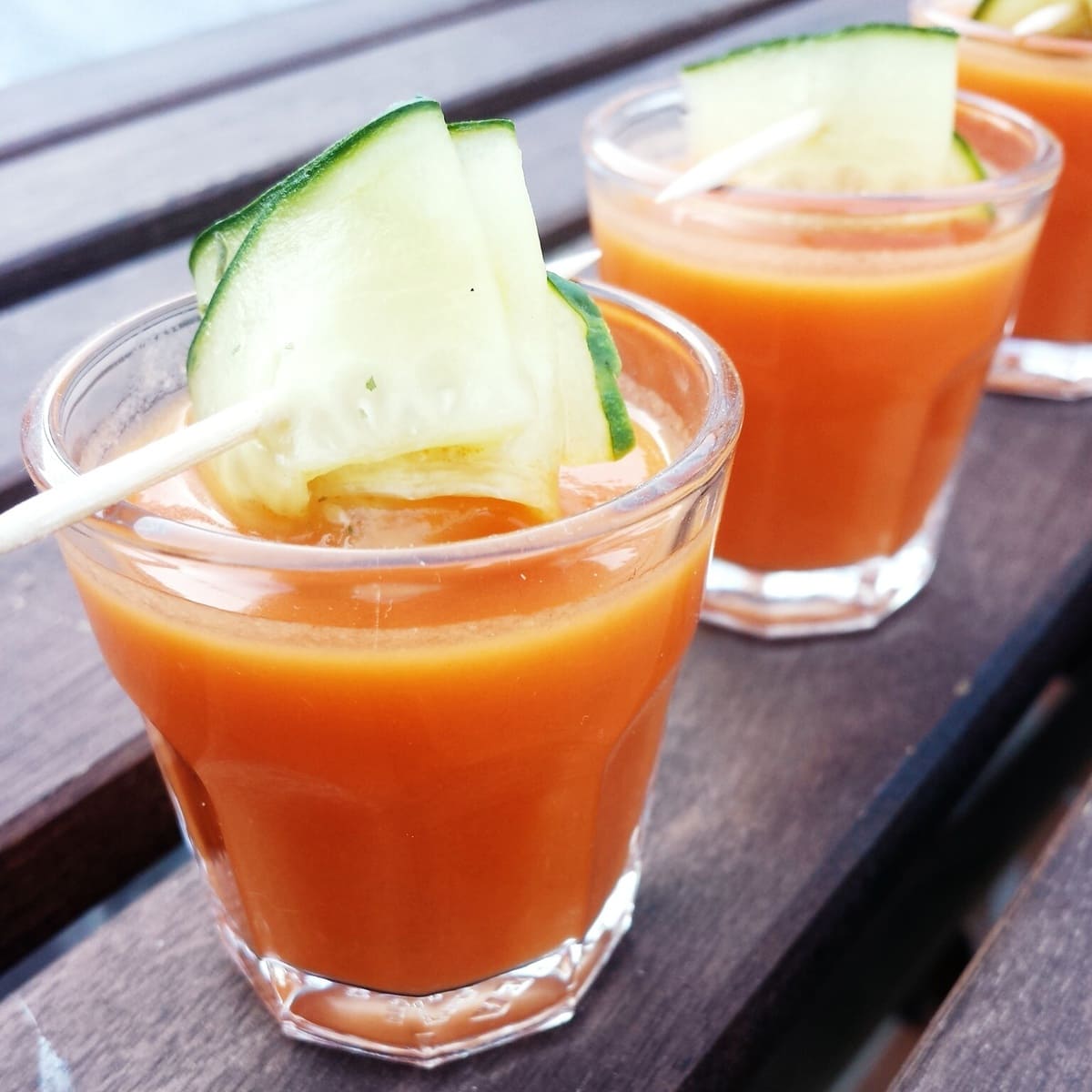 Clear glasses of gazpacho garnished with thin cucumber slices lined up on a wooden surface.