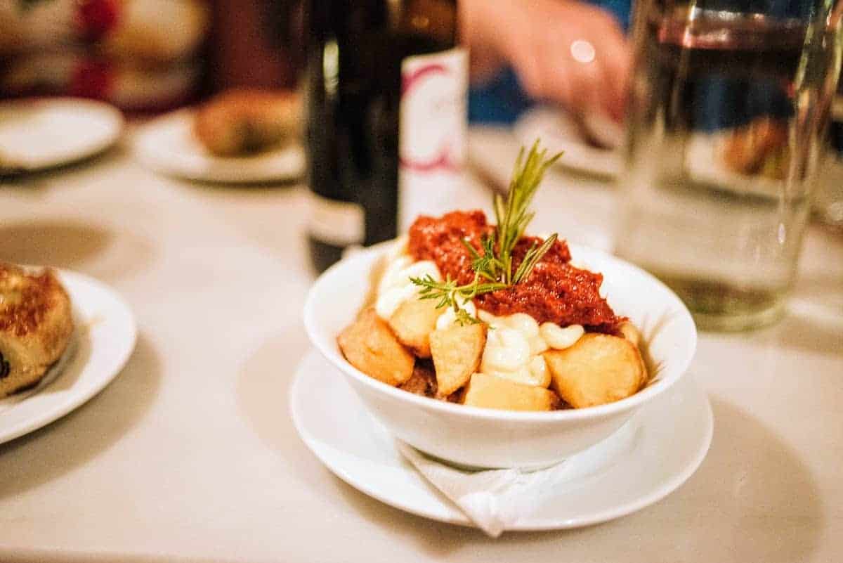 Dish of fried potato chunks topped with garlic mayonnaise and a spicy red sauce and garnished with a sprig of herb.