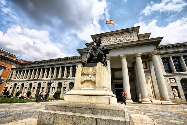 Low angle of a stone statue in front of the grand facade of the Prado Museum.