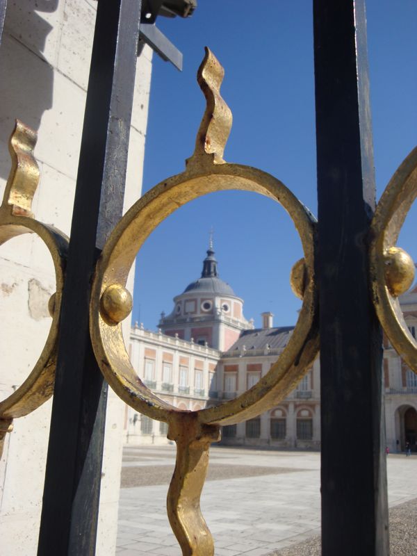 Close-up of a decorative metal gate, with the Royal Palace of Aranjuez visible through it.