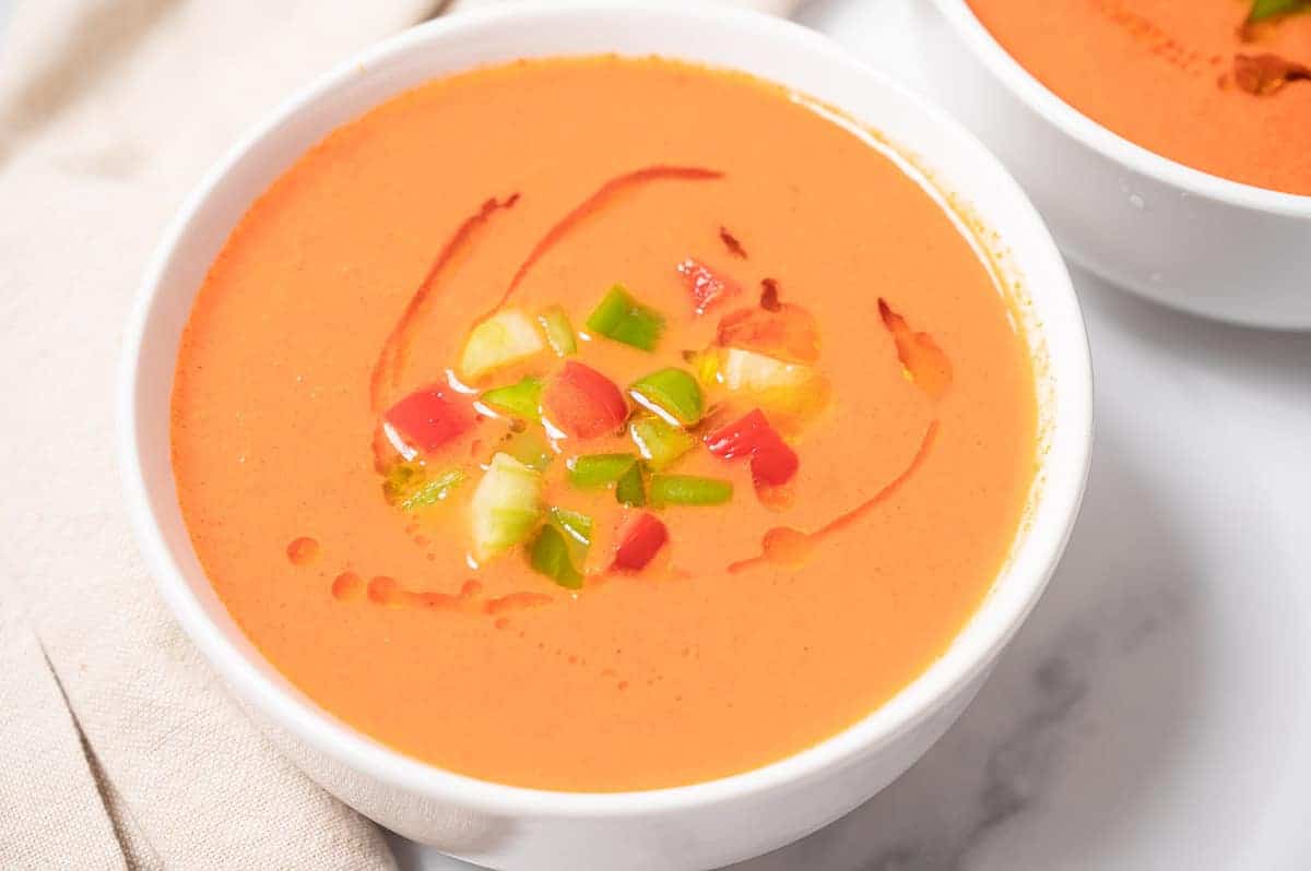 Close-up of a bowl of pale orange gazpacho topped with diced veggies and olive oil.