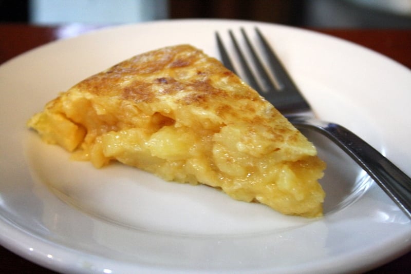 Slice of Spanish potato omelette with a gooey center, on a white plate with a fork.