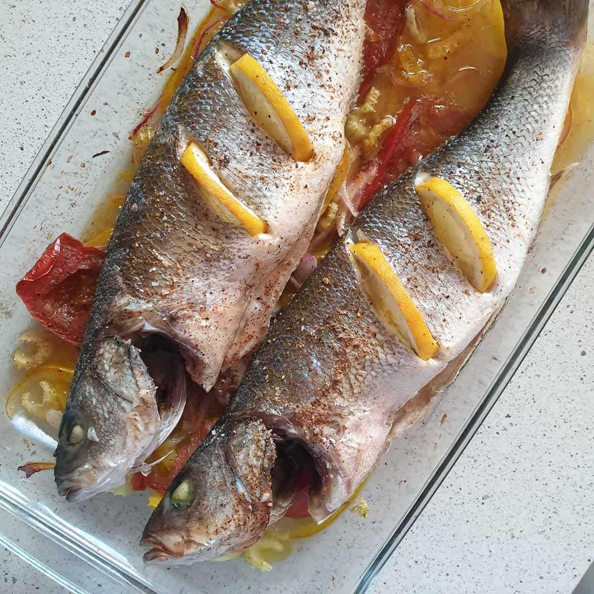 Two whole fish in a clear serving dish with sauce and sliced lemons stuck into them.