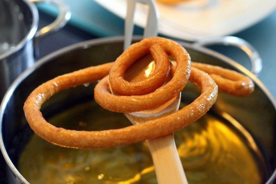 A long, thin spiral of freshly fried dough balanced on a wooden spoon over hot oil.