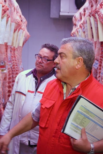 Two men discuss the pieces of raw meat around them, one of them holding a folder.