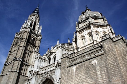 Looking up at Toledo's stately cathedral, with a tall tower and a blue sky behind it.