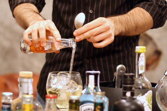 Close-up of a bartender pouring tonic into a large cocktail glass, with bottles in the foreground.