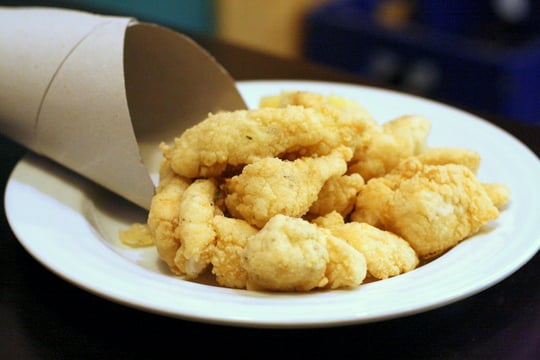 White plate with a pile of fried fish pieces spilling out of a cardboard cone.