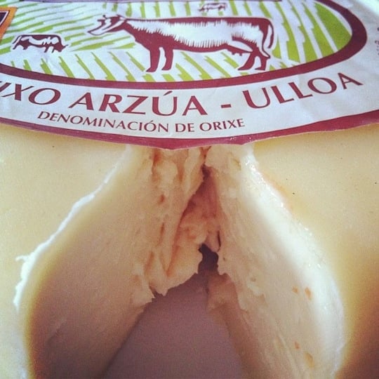 Close-up of a wheel of Arzúa-Ulloa cheese with a slice missing, revealing a creamy center.