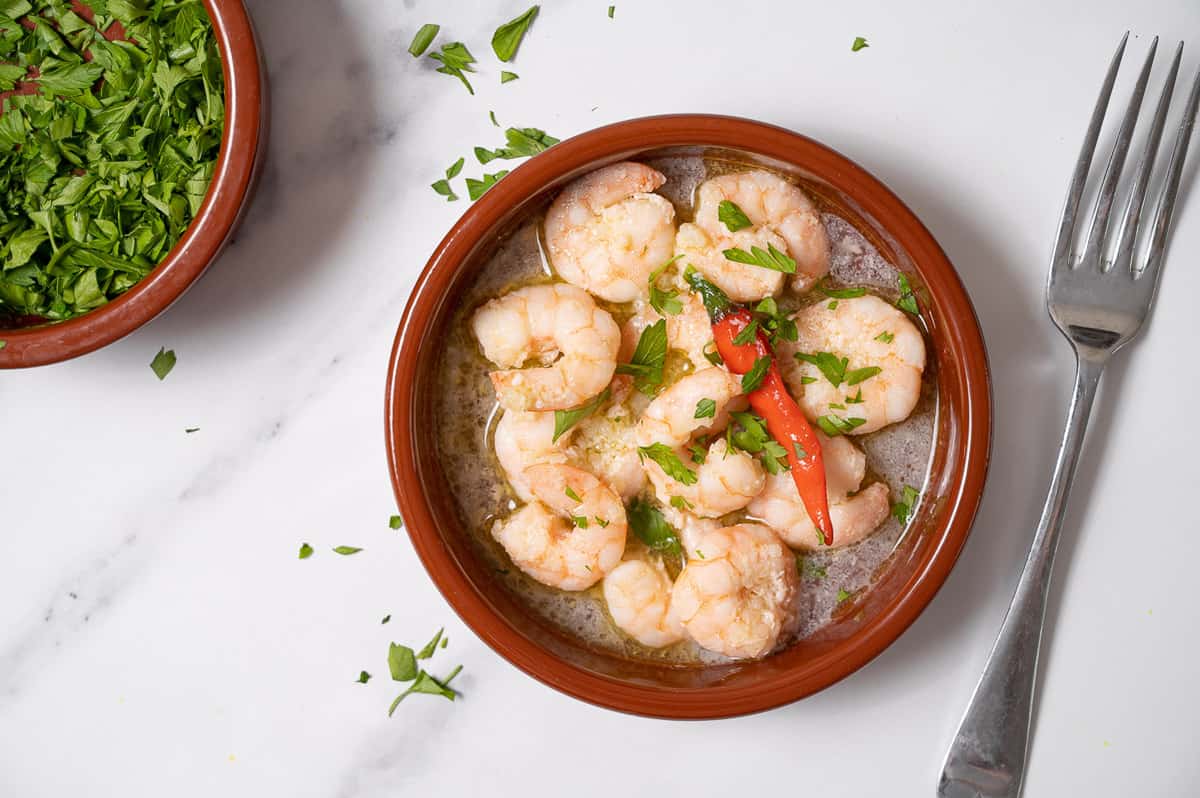 Overhead view of a clay dish of shrimp with chopped herbs and a red chili pepper. 