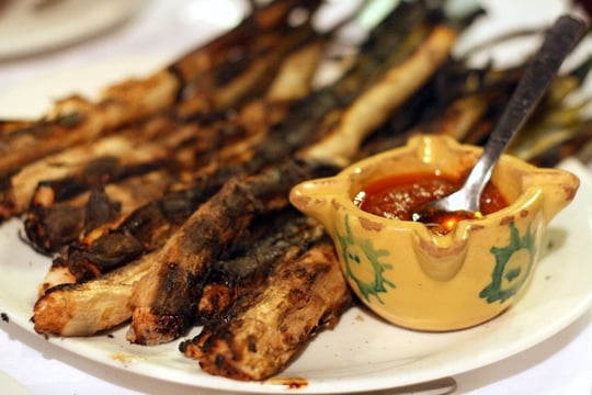 A plate of charred calçots (Catalan green onions) with a yellow bowl of bright red sauce.