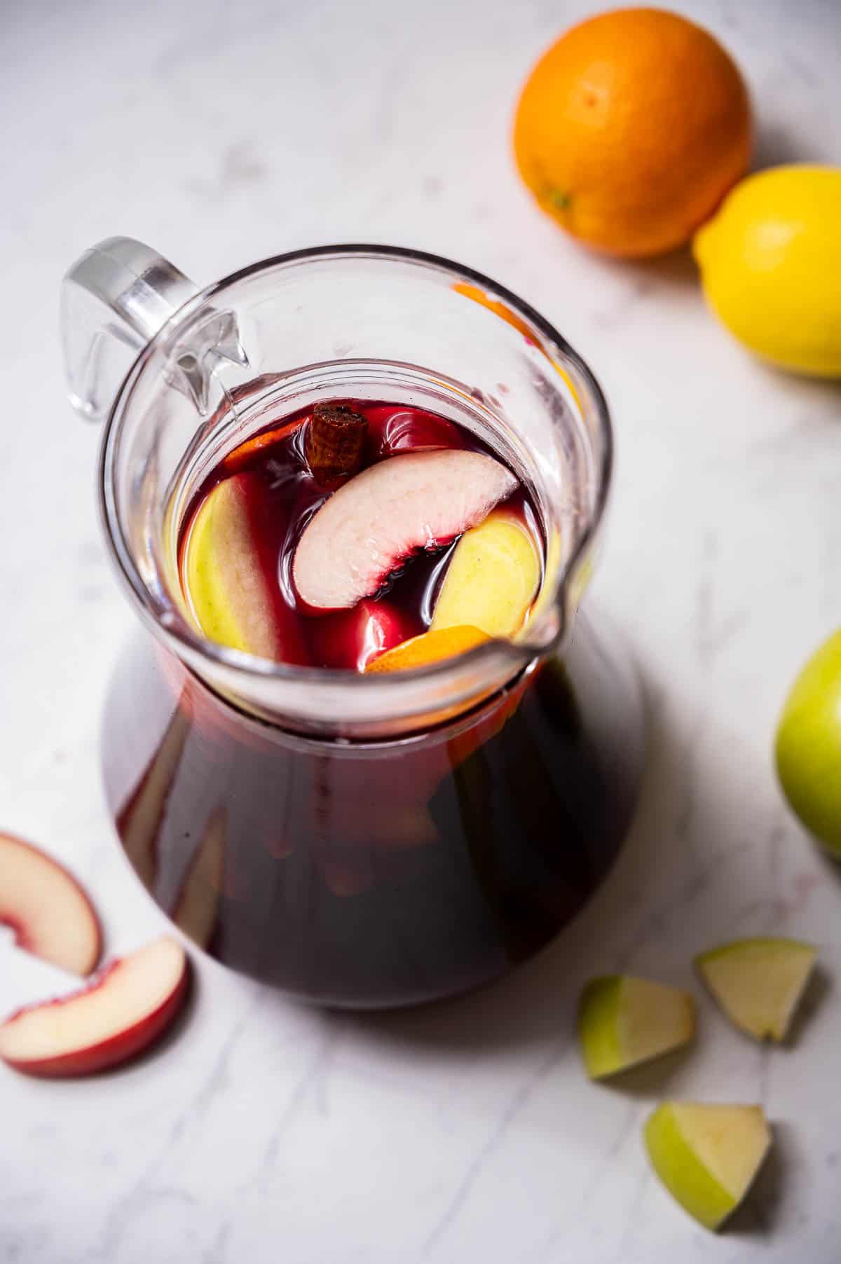 A pitcher of red wine sangria on a white countertop with fruit in the background