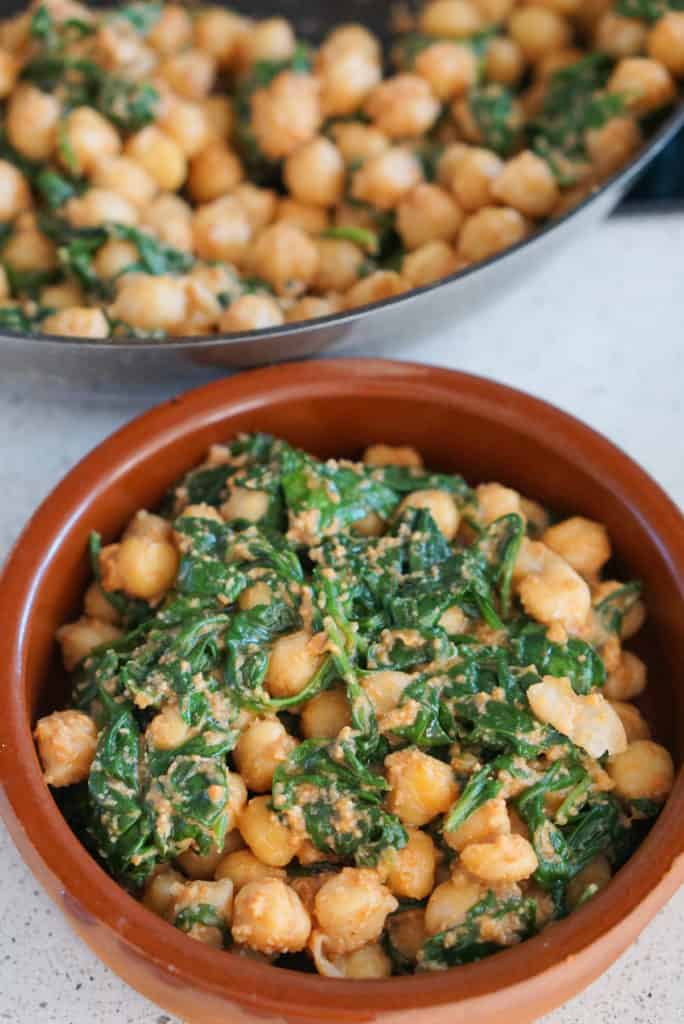 Spinach and chickpea stew in a clay dish in front of a pan full of more.