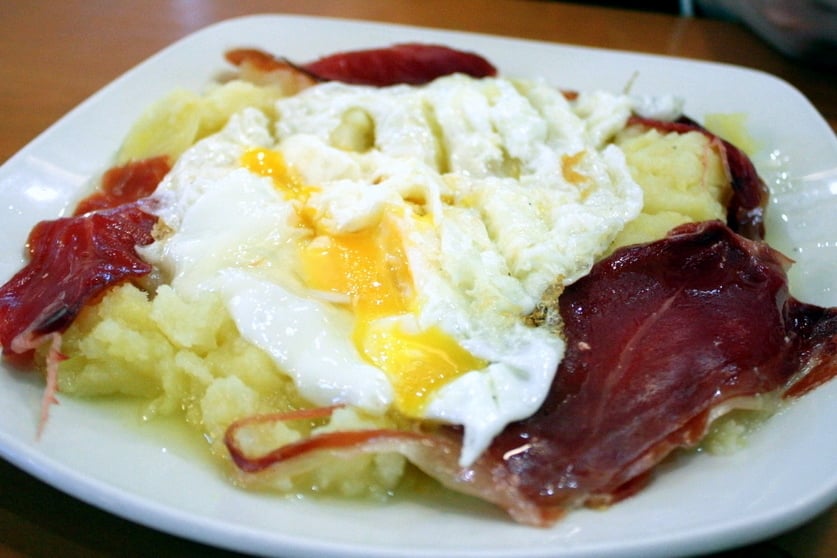 A plate of potatoes with cured ham and runny fried eggs on top.