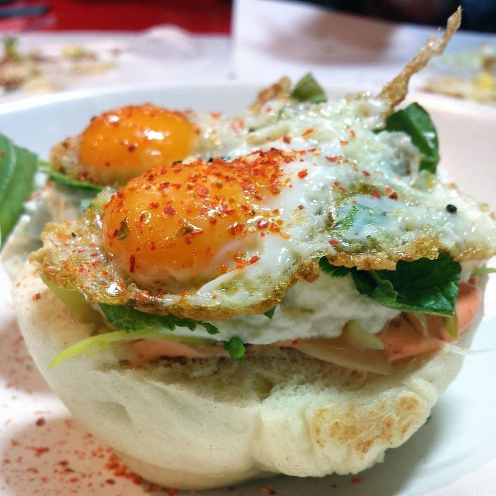 Close-up of an open-sided bao bun with fried eggs, spinach, and other toppings.