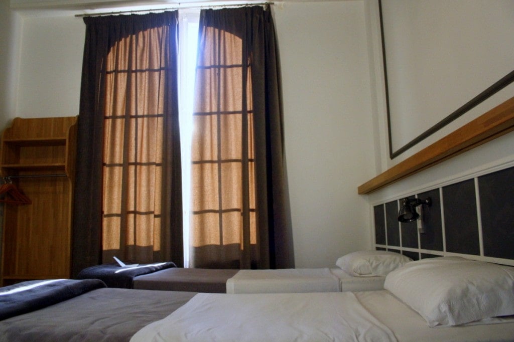 A simple hotel room with white walls, two twin beds, and sunlight filtering through brown curtains.