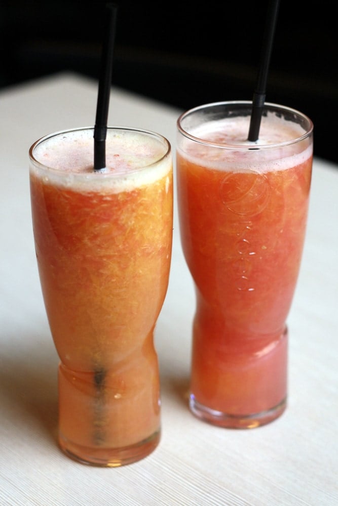 Two tall glasses of fresh juice, both shades of light reddish-orange, with black straws.