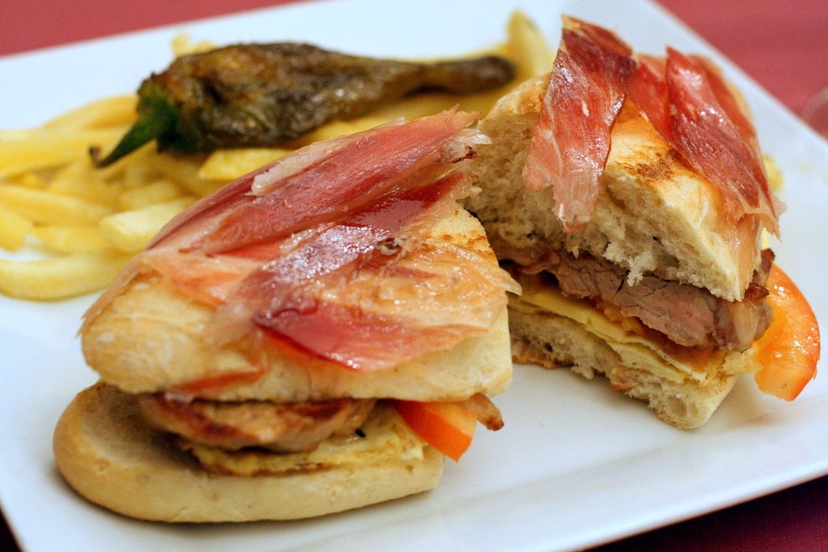 Small sandwich of pork loin, tomato, and cured ham served alongside french fries and a fried green pepper.