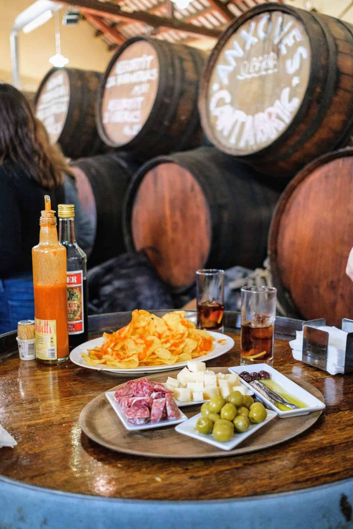 Tapas spread of cured meat, olives, cheese, anchovies, potato chips, and vermouth atop a wooden barrel.