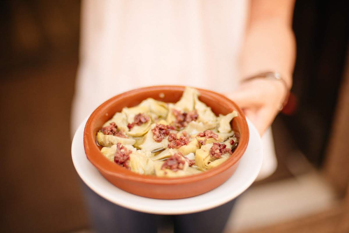 Someone holding a clay dish of artichoke hearts with diced Iberian ham on top.