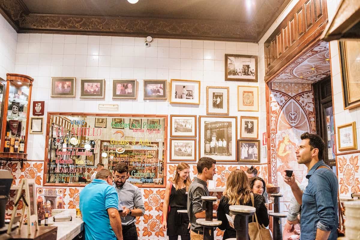 Colorful interior of a tapas bar with people standing at high tables drinking red wine.