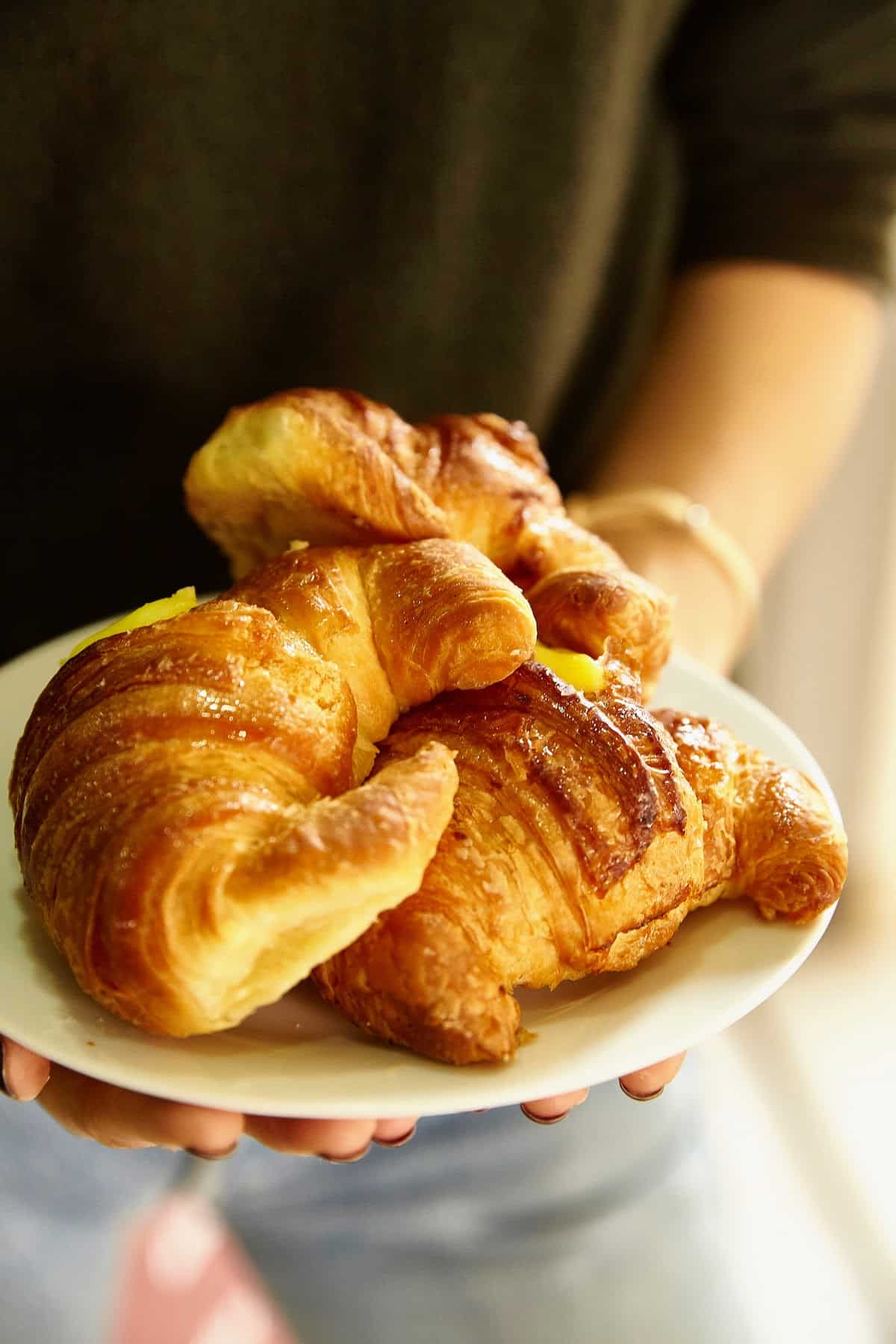 Close-up of a person holding a white plate with three large, shiny croissants.