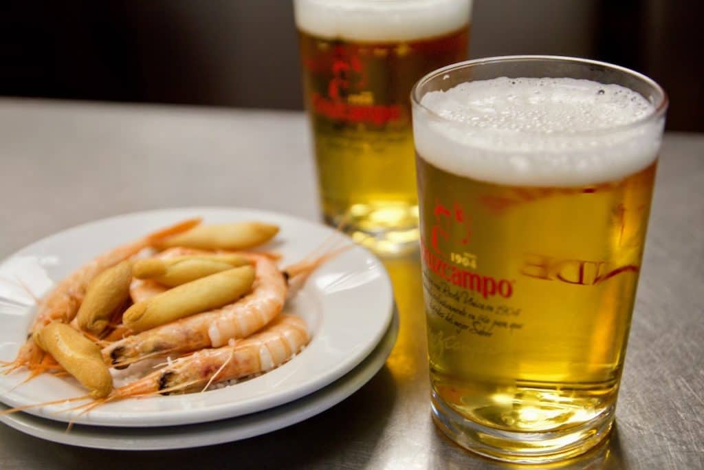 Two small draft beers next to a tapa of boiled shrimp and crunchy breadsticks.