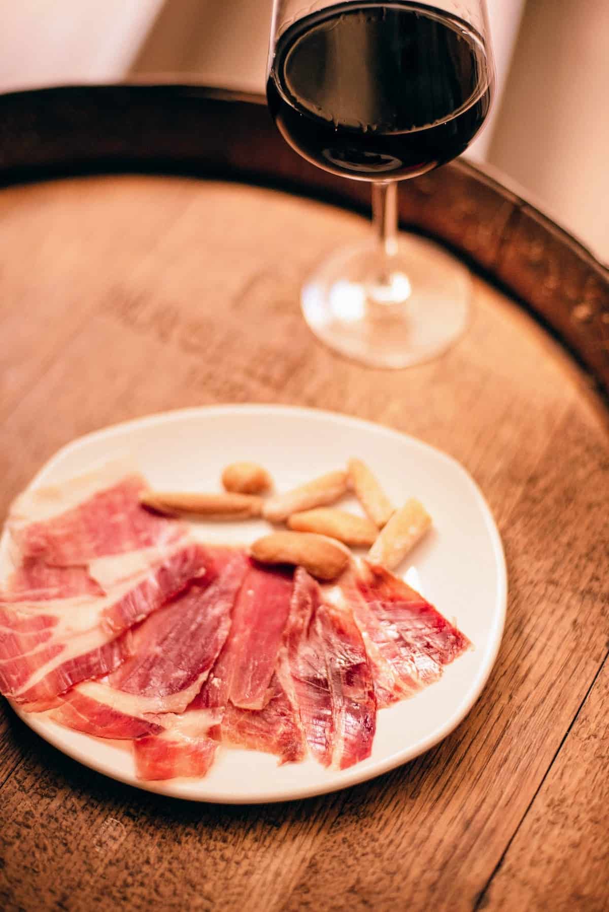 Plate of cured ham and small breadsticks beside a glass of red wine on a wooden barrel.