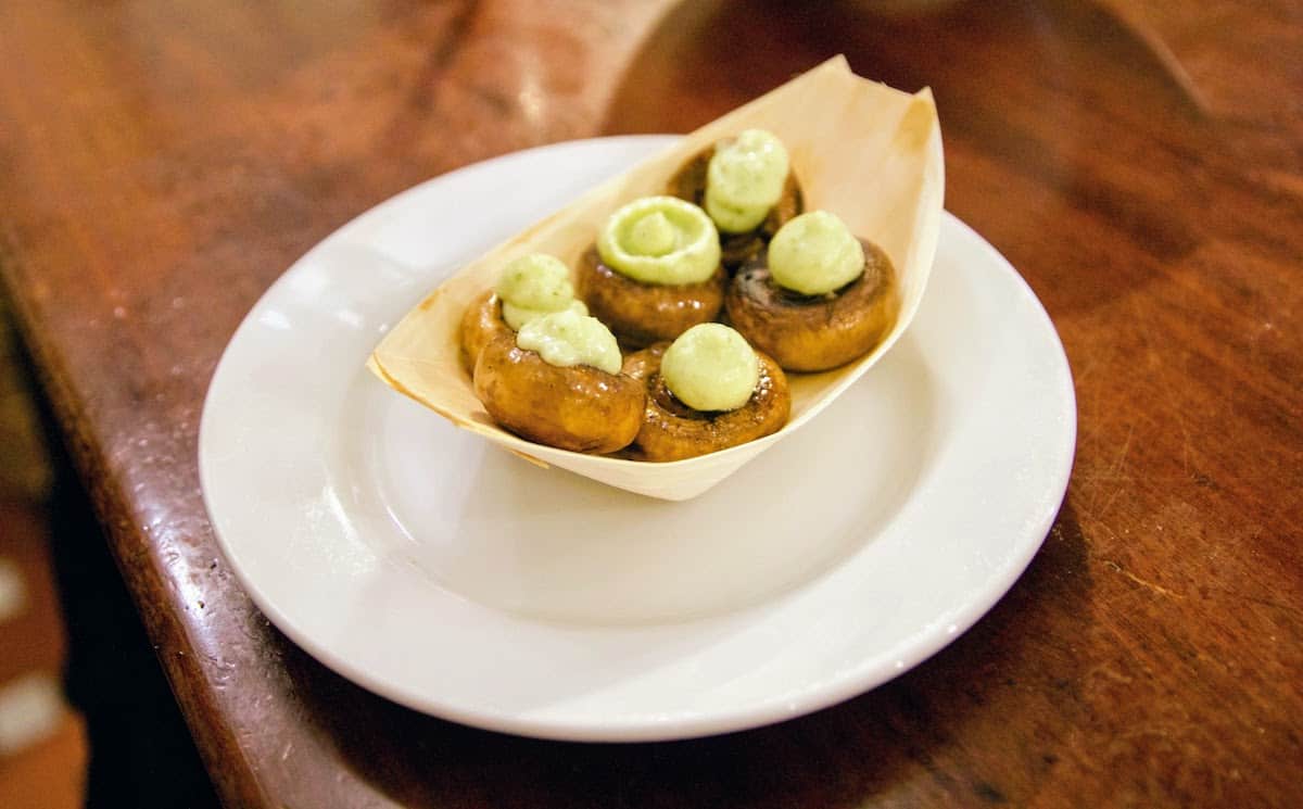 Tapas portion of six mushrooms filled with light green parsley aioli on a wooden table.