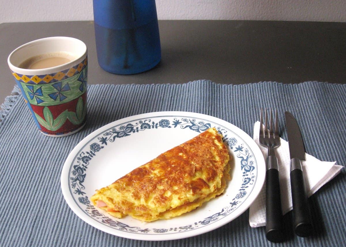 Omelet on a white plate on a table beside a fork, knife, napkin and a cup of coffee