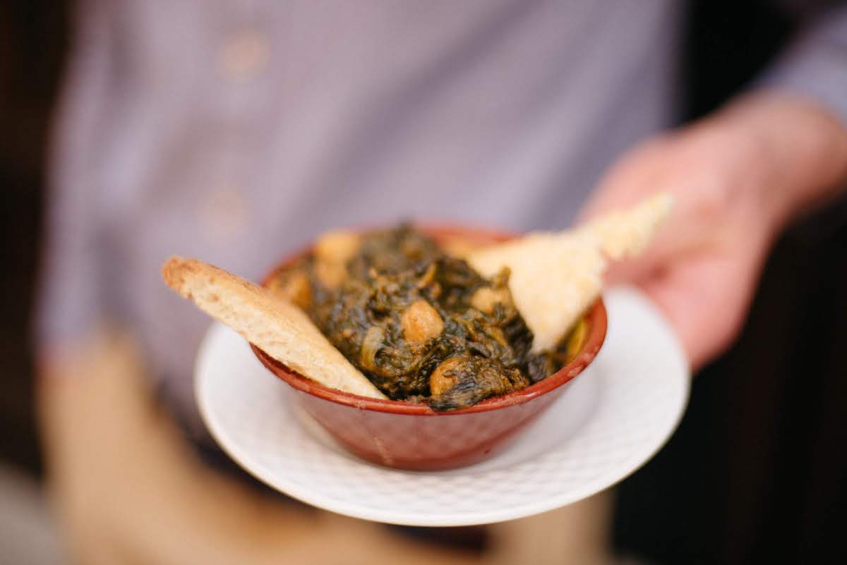 Someone holding a small clay dish of spinach and chickpea stew served with crackers.