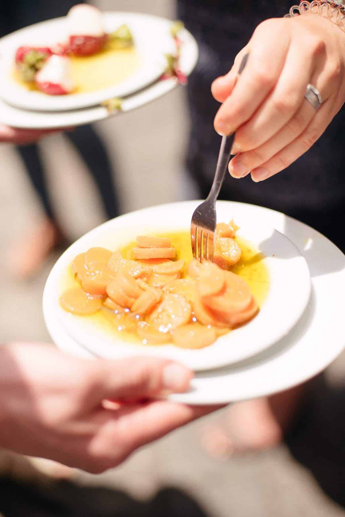 A person's hand using a fork to eat marinated carrots in olive oil off a plate.