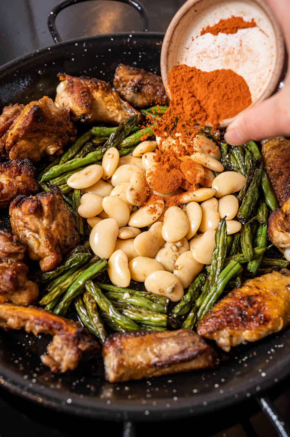 Close-up of a person's hand pouring orange paprika into a pot of meat, veggies, and beans.