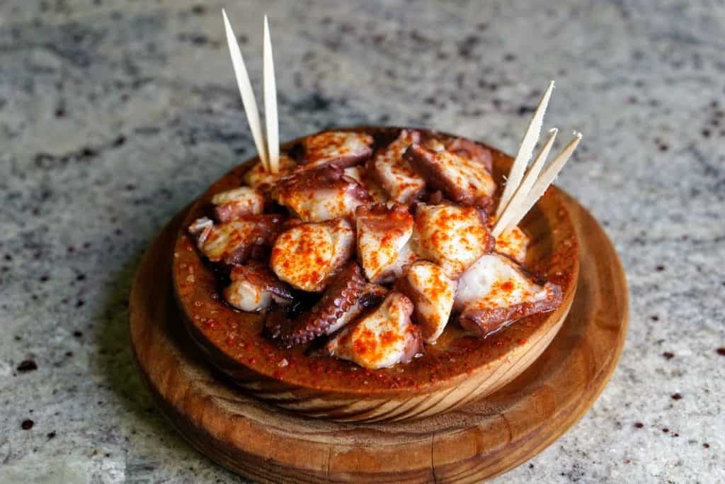 Slices of boiled octopus covered in paprika served in a clay dish with toothpicks.