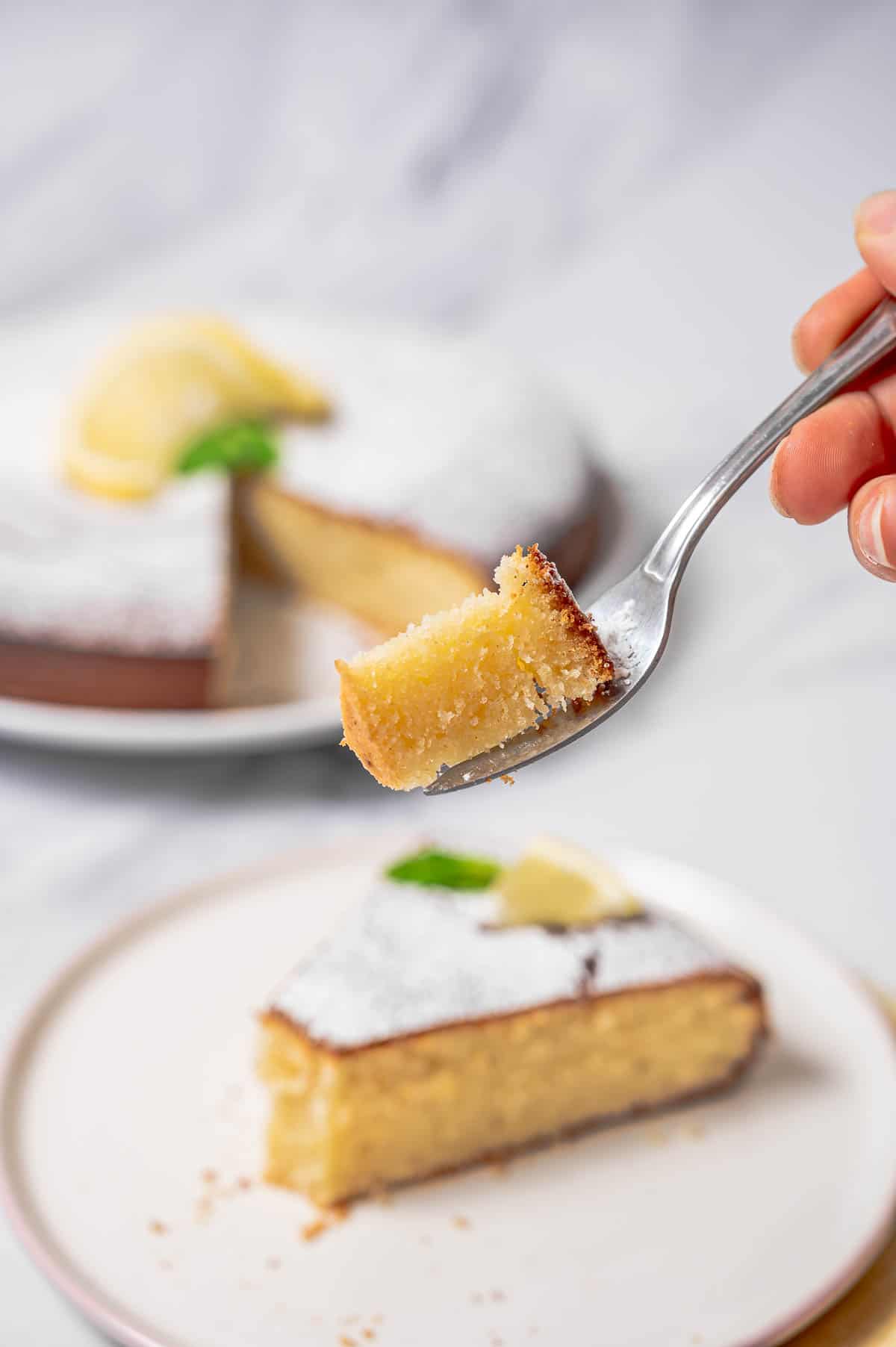 Close up of a bite of lemon olive oil cake on a fork with the rest of the cake in the background.