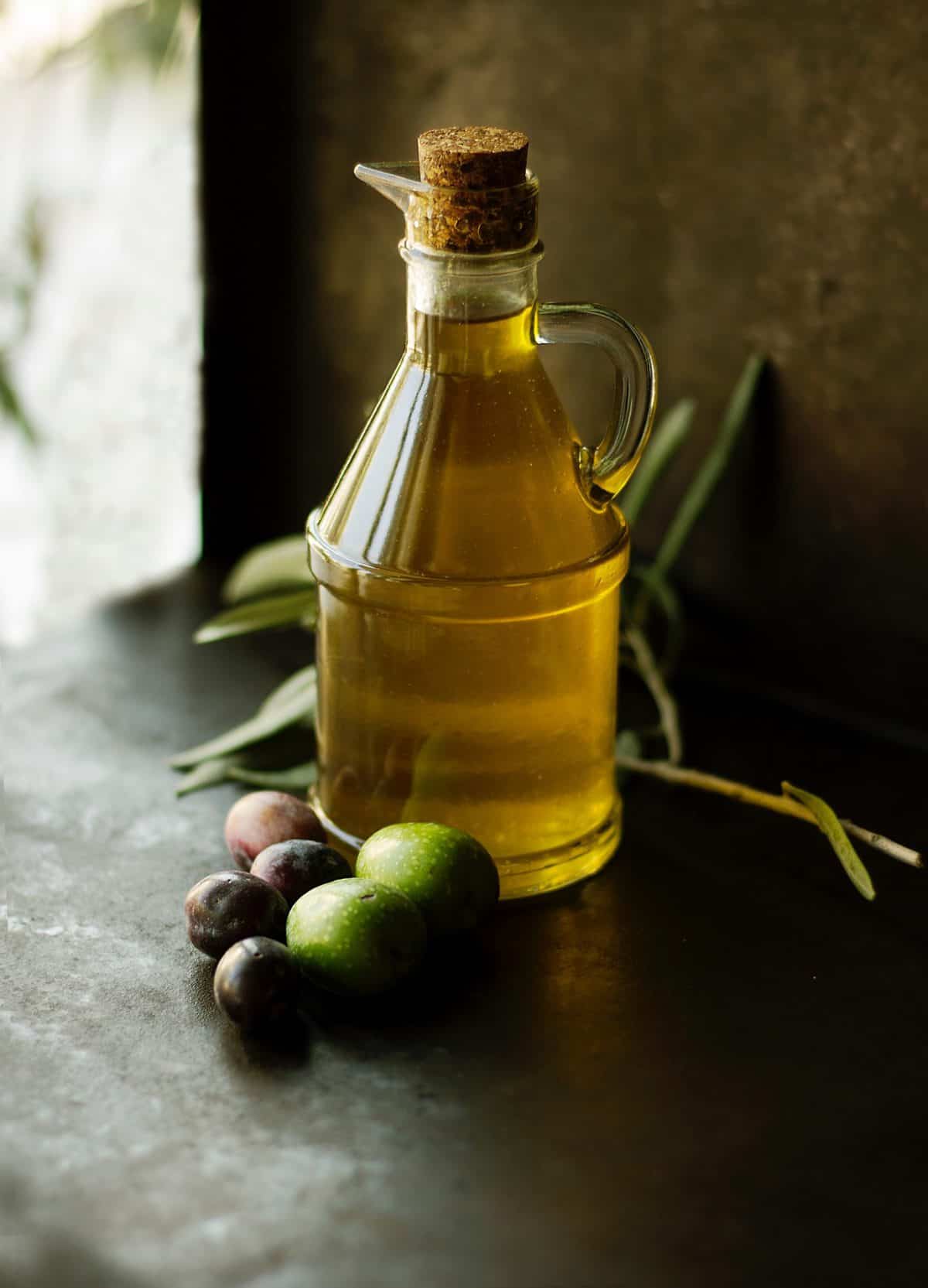 Clear bottle of olive oil with a spout and cork stopper beside a handful of olives.