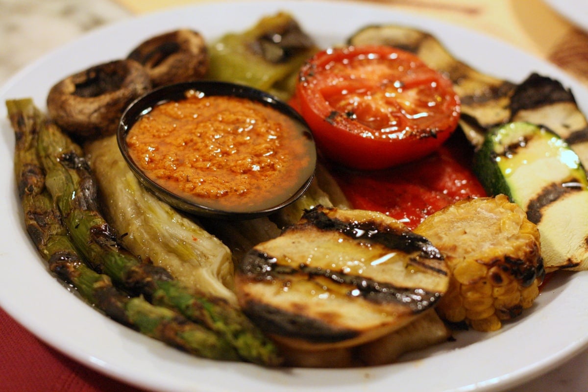 Mediterranean roast vegetables on a white plate with a small bowl of romesco sauce in the middle.