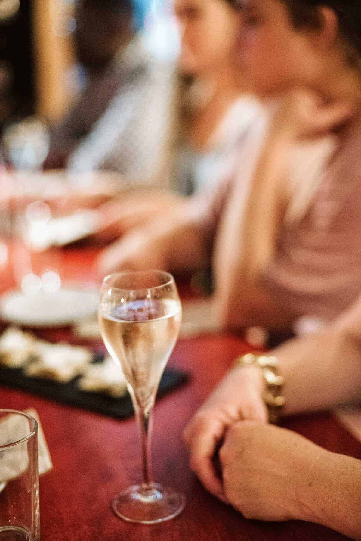 A glass of cava on a red tabletop with food and people in the background.