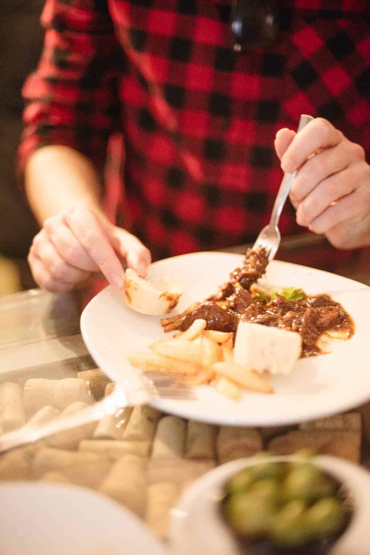 Close up of a person eating a meat dish while holding a small chunk of bread in the other hand.