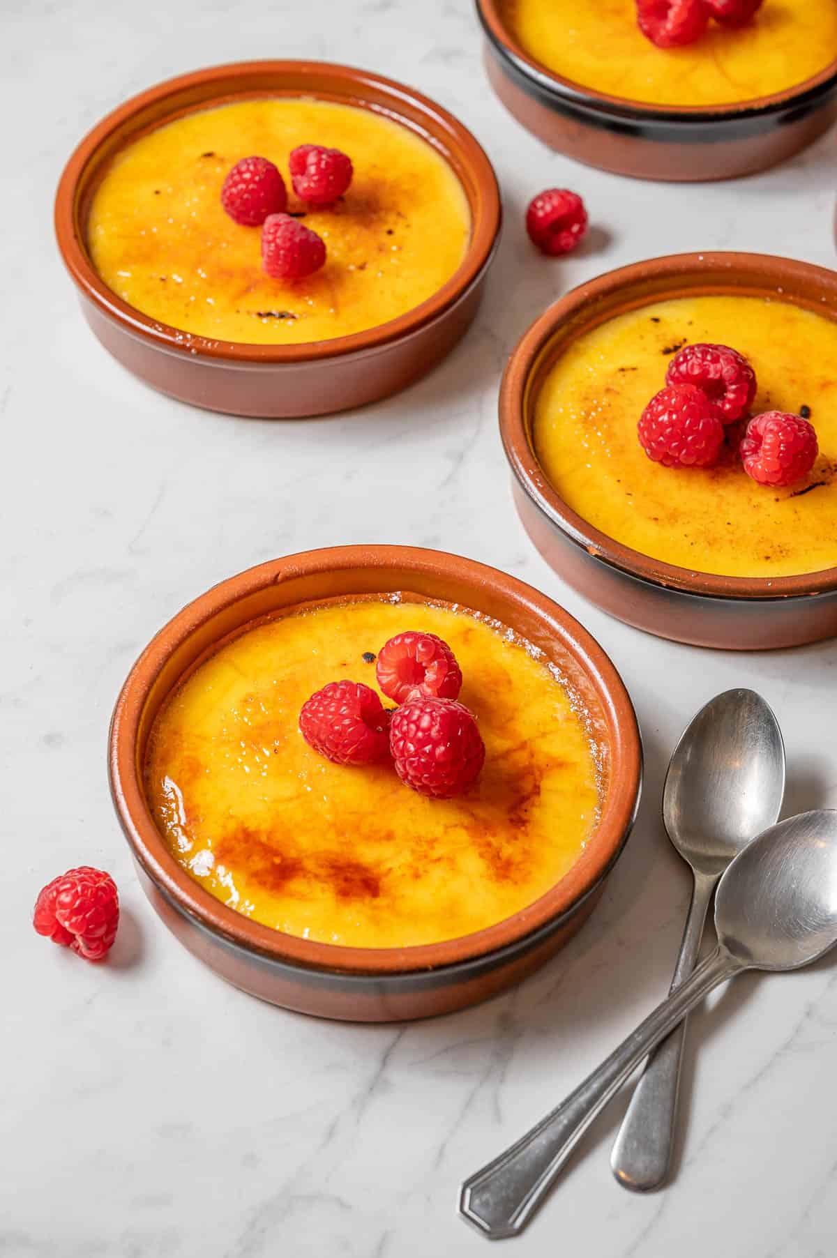 Four individual crema catalanas in clay dishes with raspberries.