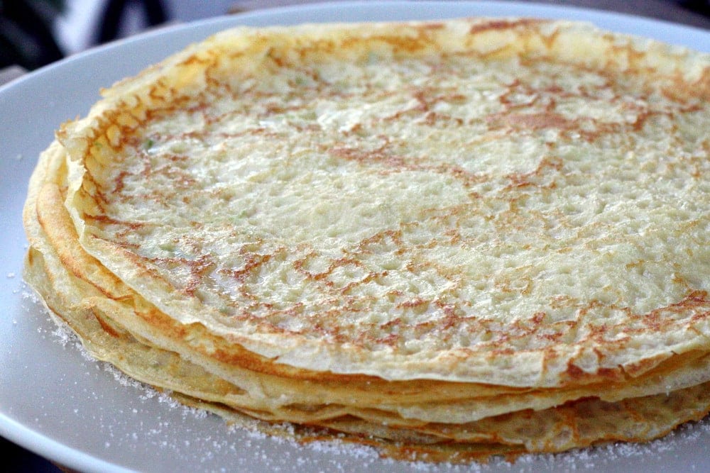 A stack of thin frisuelos, or Spanish crepes, on a white plate with powdered sugar.