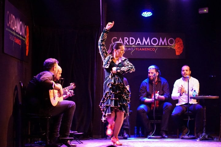 A flamenco dancer performs onstage with four men playing guitars and clapping