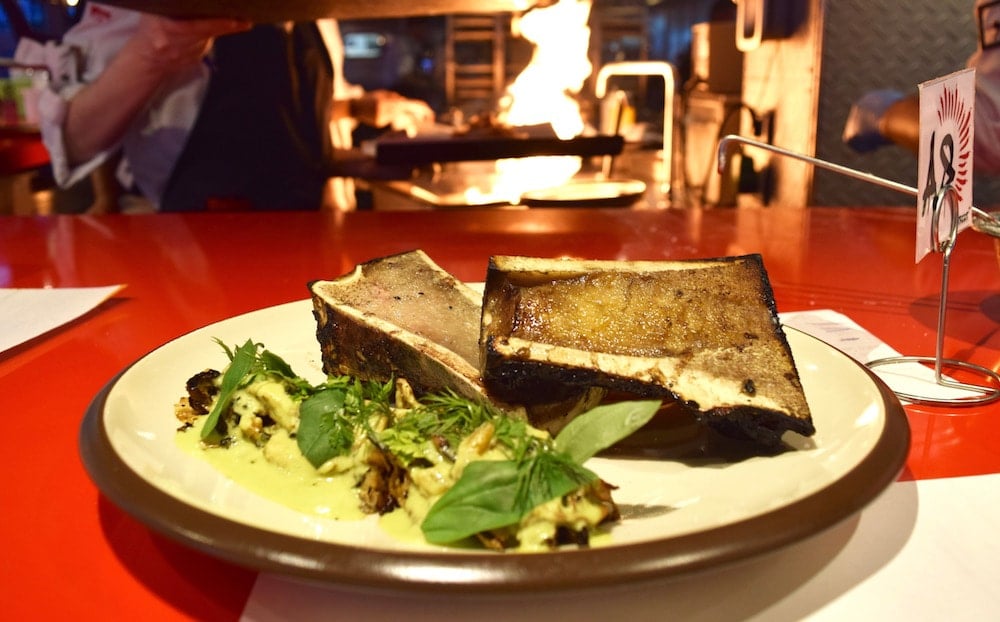 Close-up of a plate of grilled bone marrow, churros, and green curry on a red table